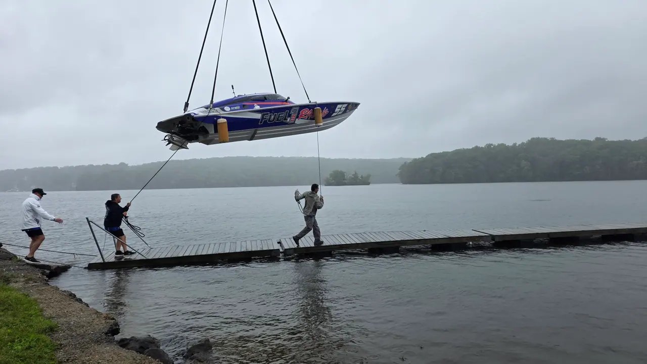Crew on a lakeside dock steady the #55 hull as a crane lowers it toward Lake Hopatcong