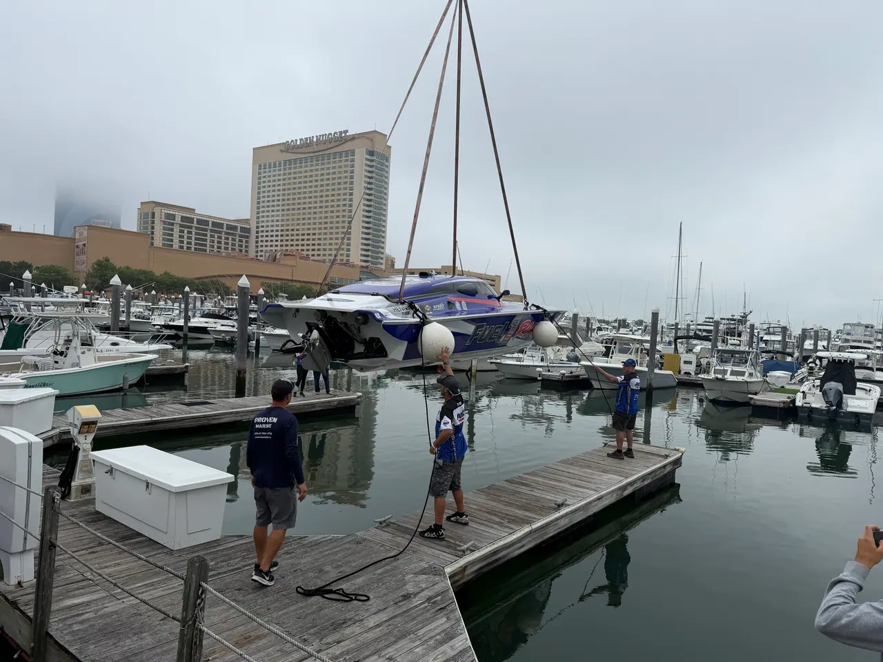 Crew on a marina dock guide a Fuel 1 race boat down from a crane, hotel towers in the fog