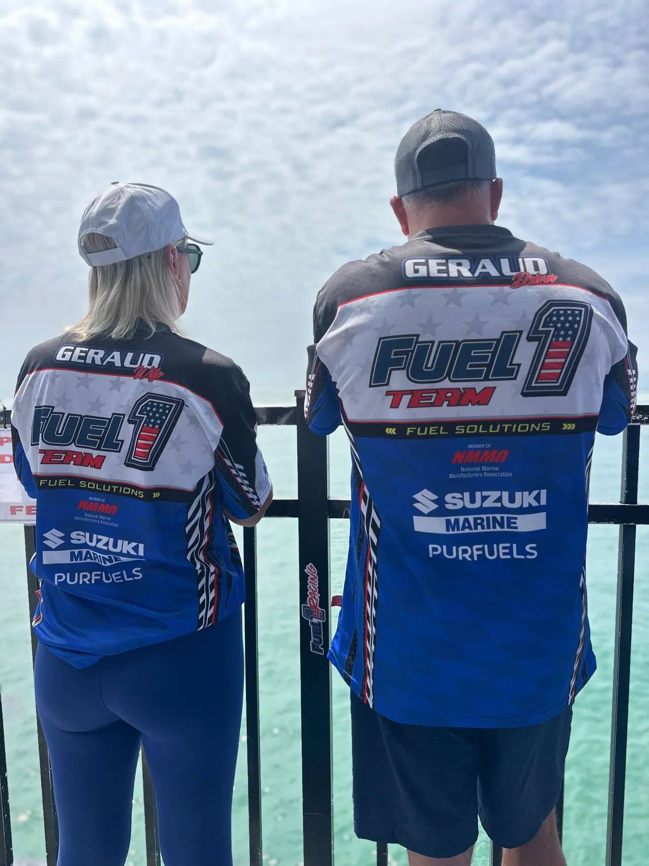Two team members seen from behind at a waterfront railing, both jerseys printed GERAUD with Fuel 1 Team, Suzuki Marine and PurFuels logos