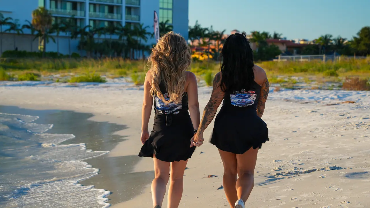 Two women in Fuel 1 outfits walking hand in hand along the shoreline at Clearwater